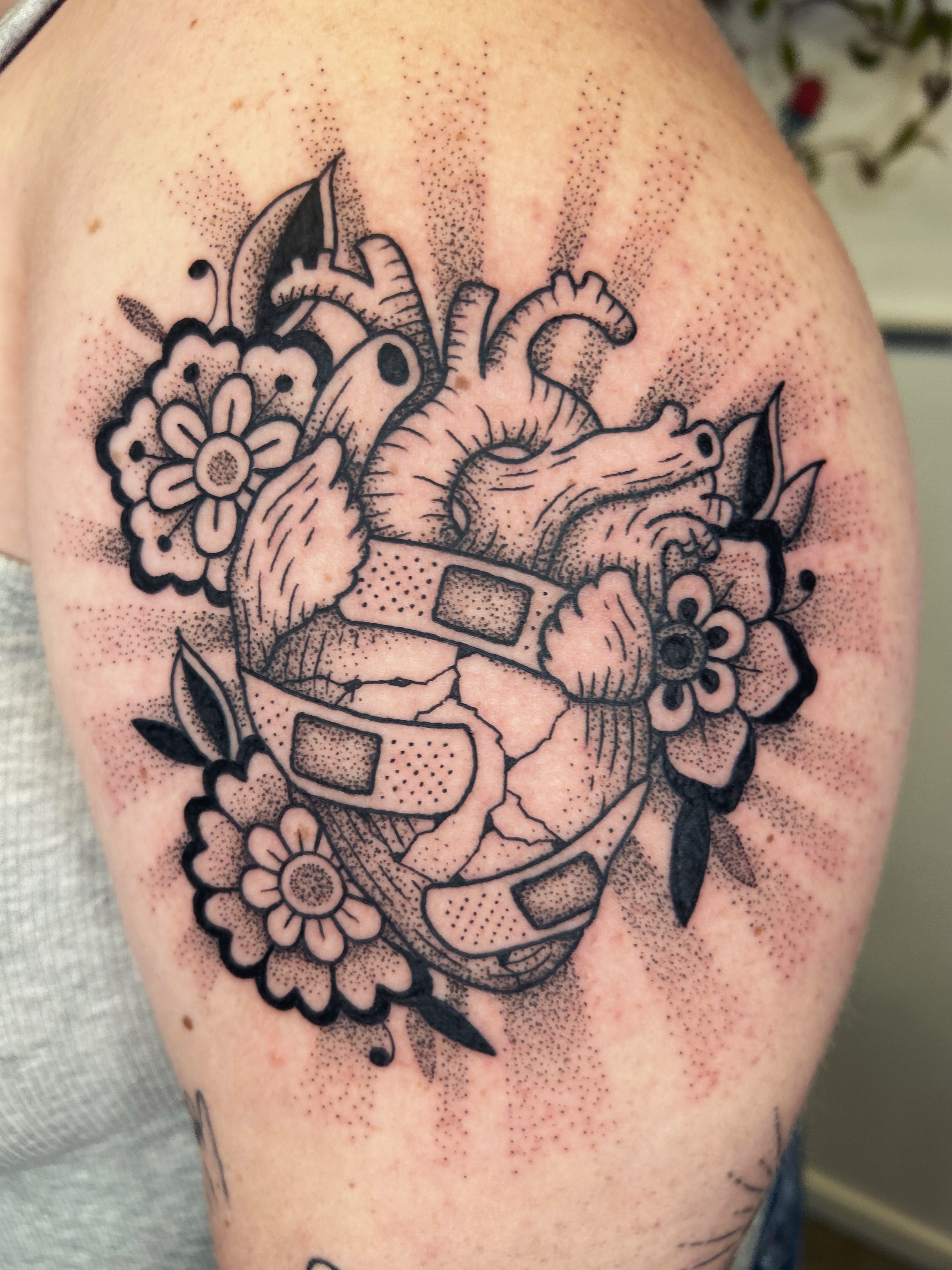 Tatouage coeur anatomique entoure de fleurs et de pansements sur l'epaule, dotwork noir et gris